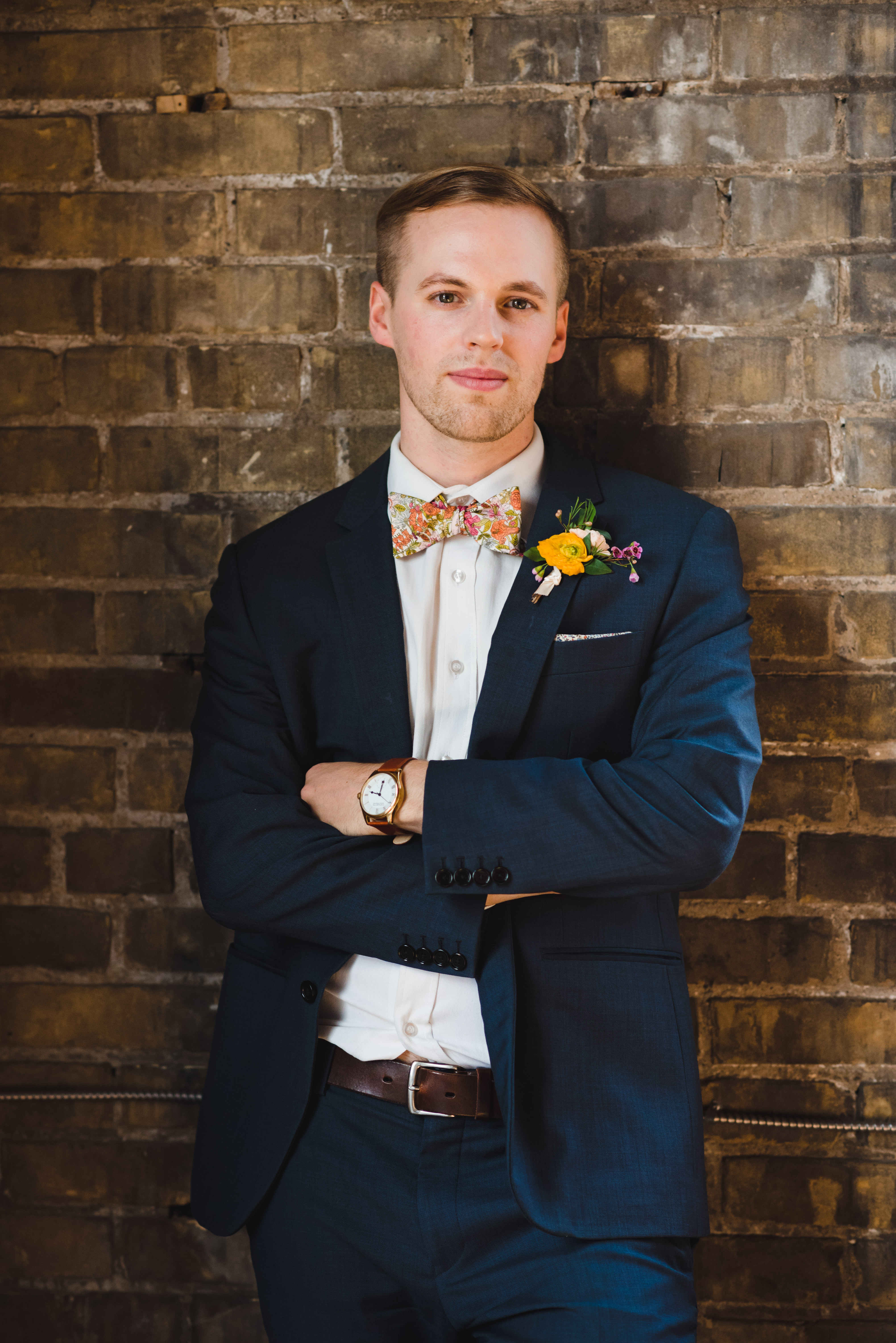 closeup of a groom wearing a blue suit with floral bowtie and boutonniere while leaning against old brick wall of the Jam Factory Toronto wedding photography