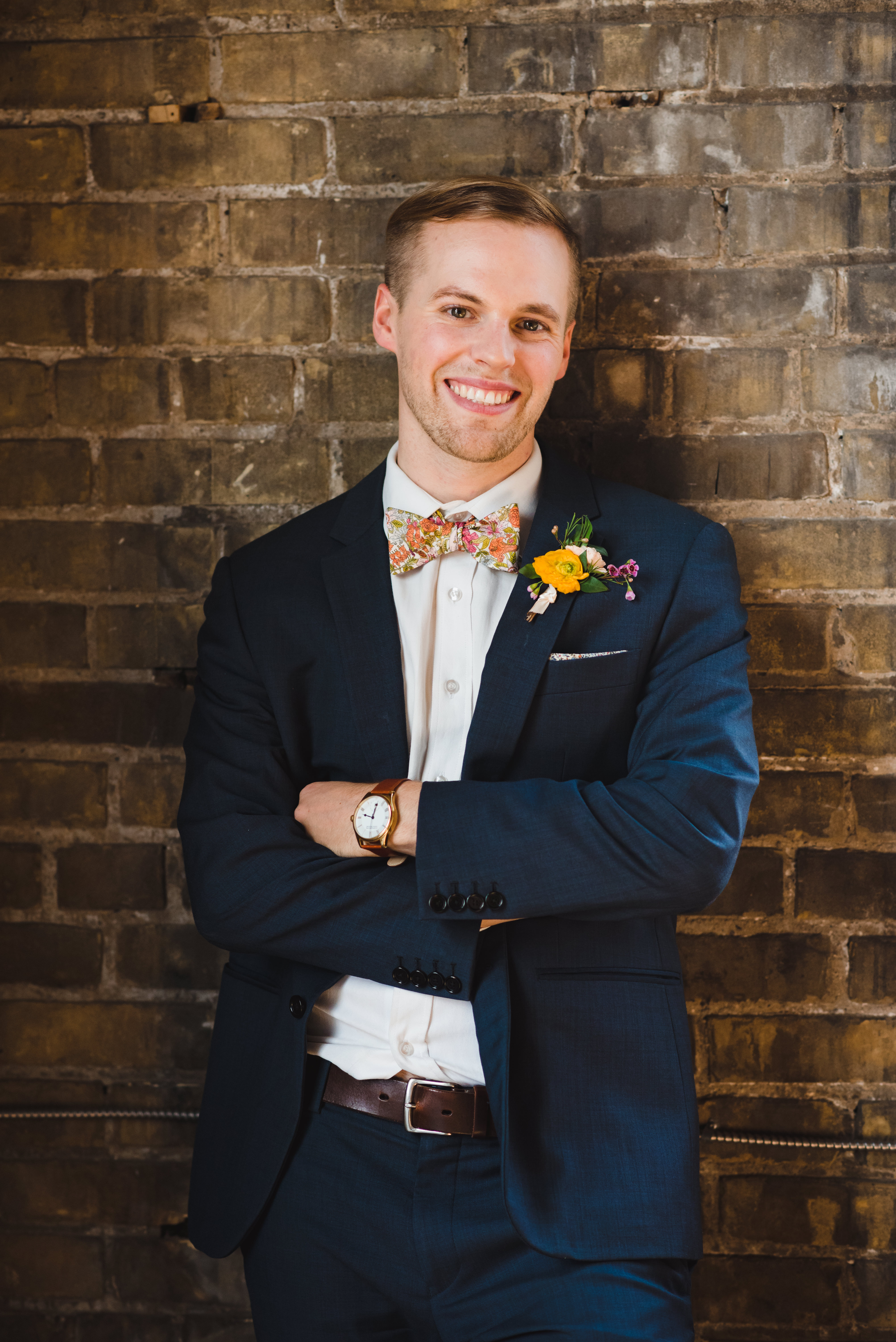 smiling groom wearing a blue suit with floral bowtie and boutonniere while leaning against old brick wall of the Jam Factory Toronto wedding photography