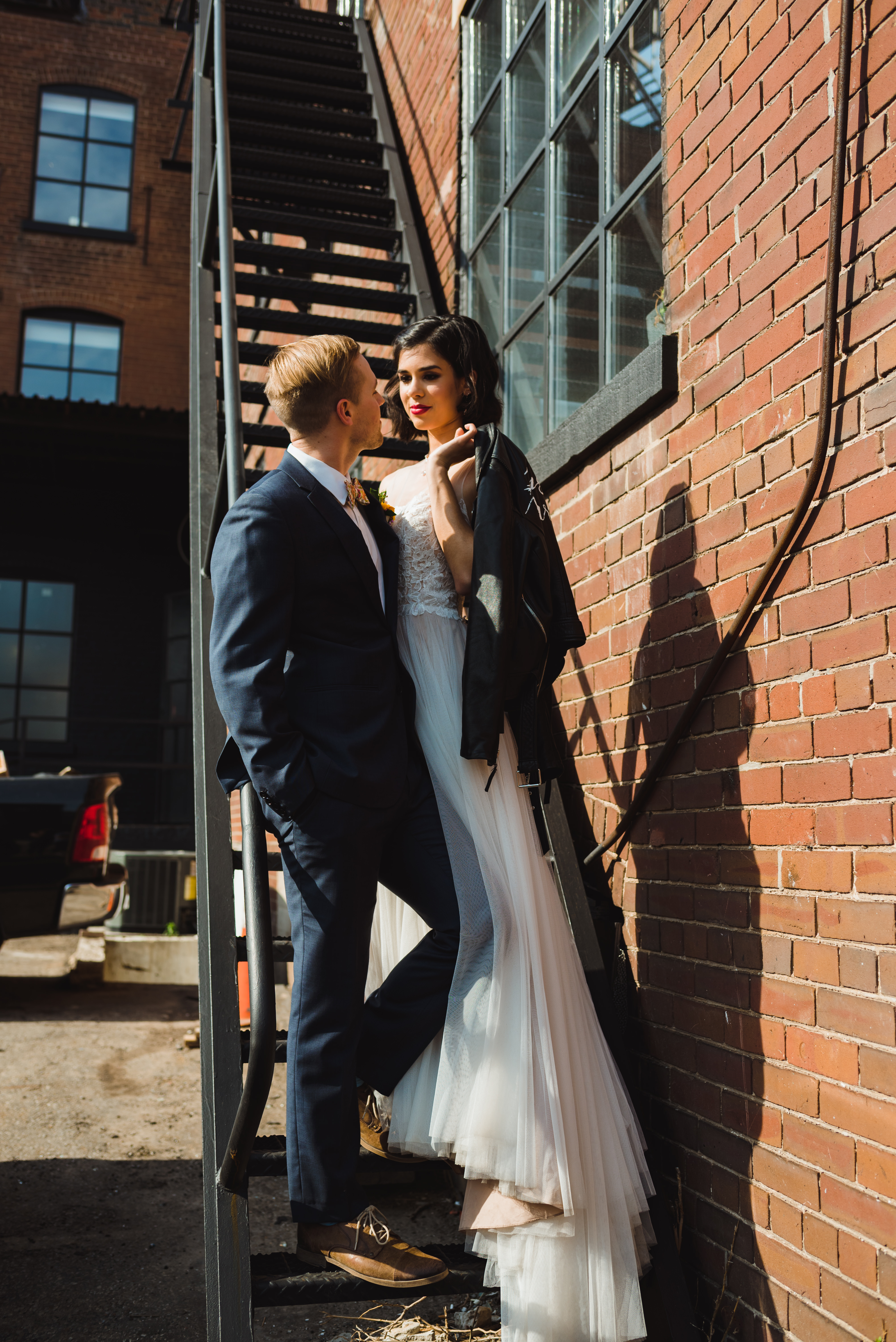 bride holding just married leather jacket over her shoulder and standing next to her groom in loving embrace on black fire escape staircase of the Jam Factory Toronto wedding photography