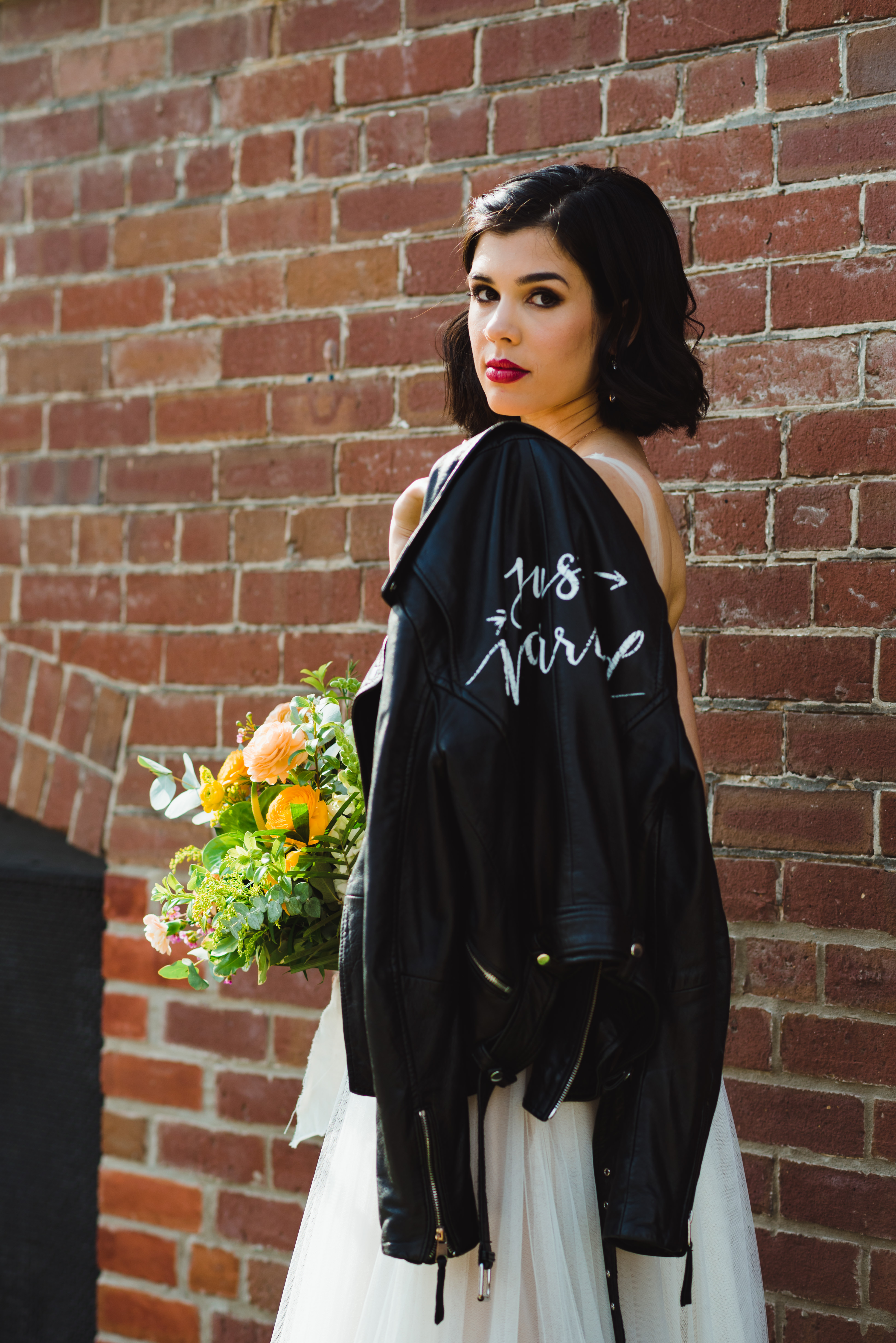 bride with just married leather jacket over her shoulder and holding bouquet standing in front of red brick wall of the Jam Factory Toronto wedding photography