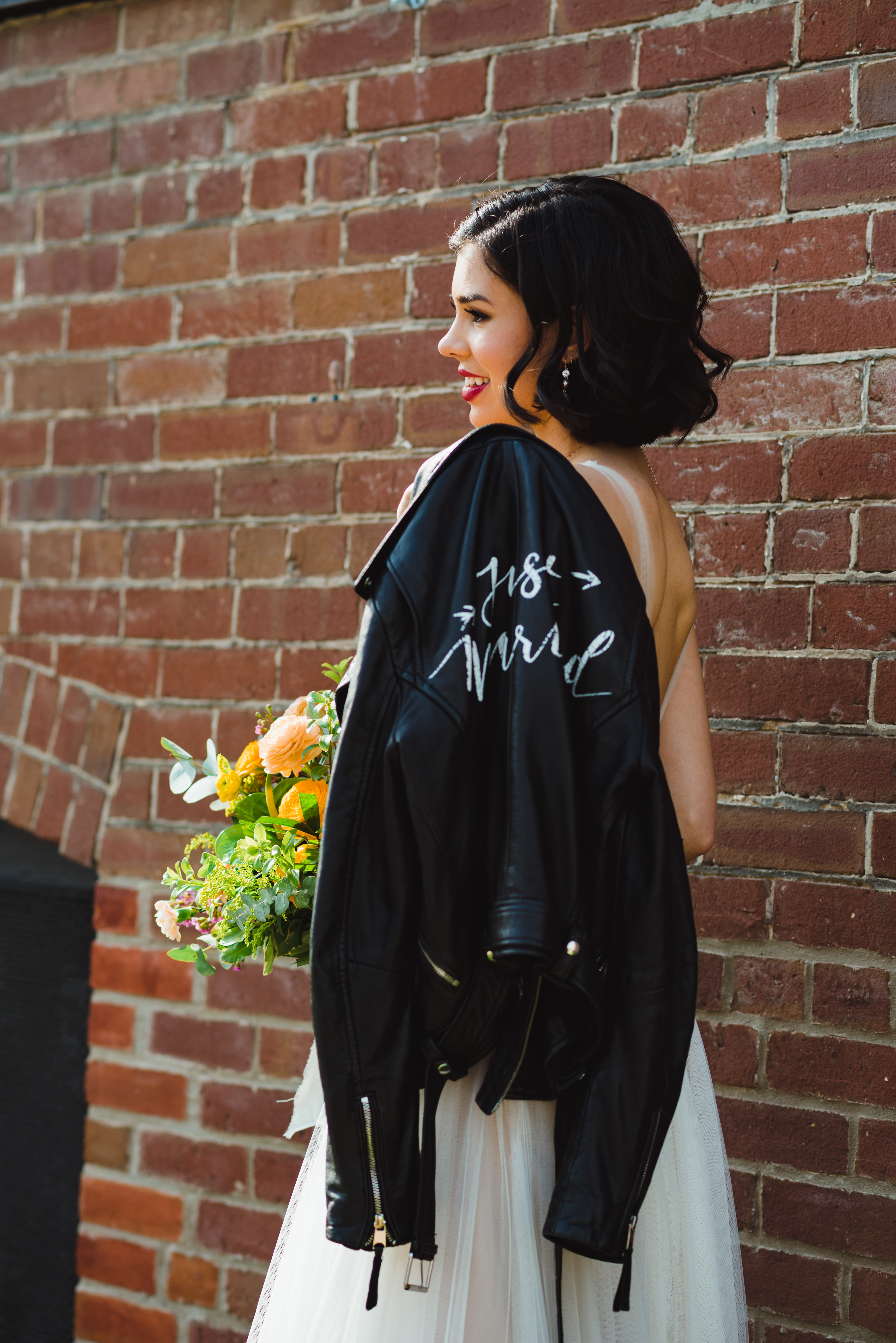 bride with just married leather jacket over her shoulder and holding bouquet standing in front of brick wall of the Jam Factory Toronto wedding photography