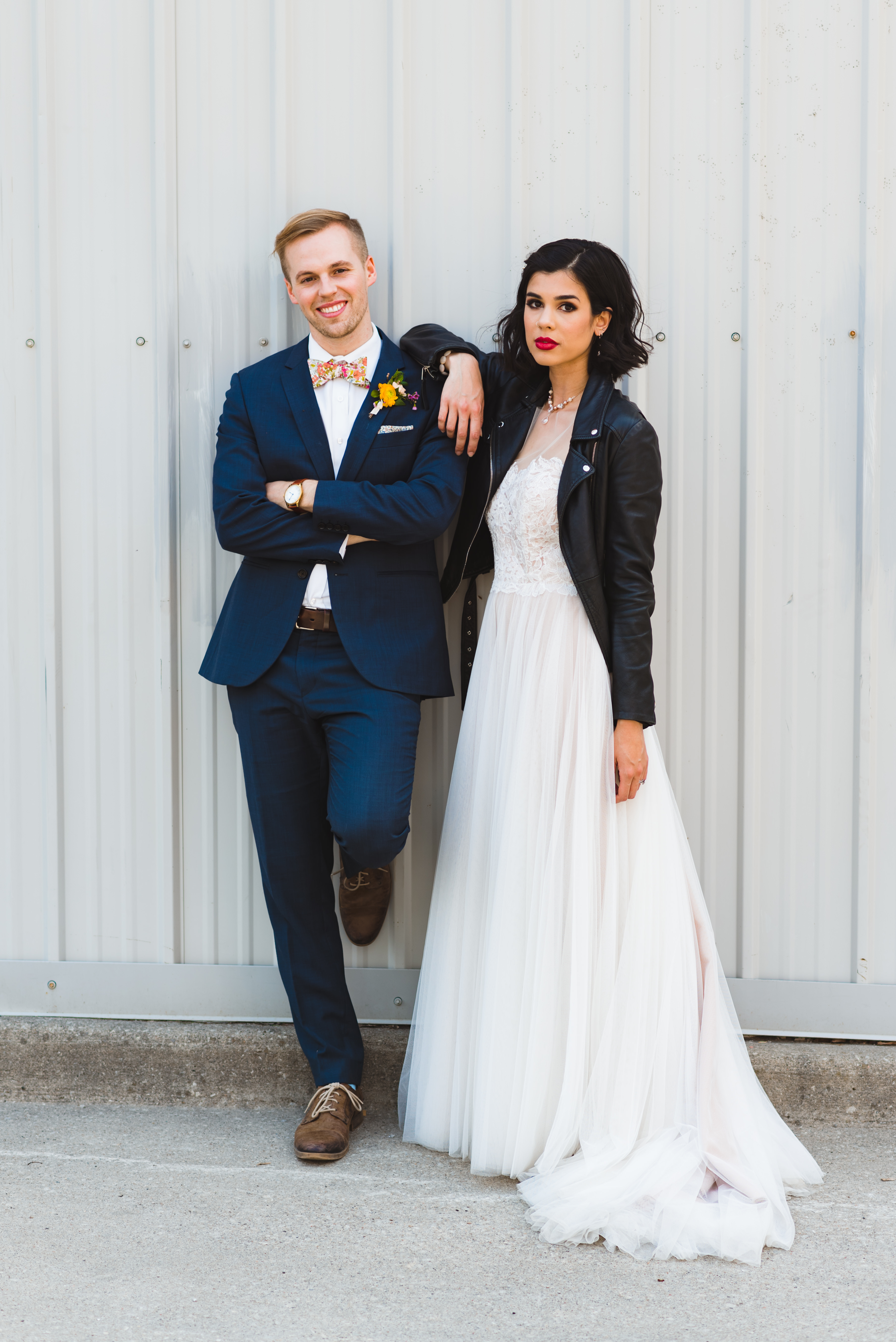 bride wearing black just married leather jacket over white wedding dress with one arm on grooms shoulder in front of white metal wall Toronto wedding photography