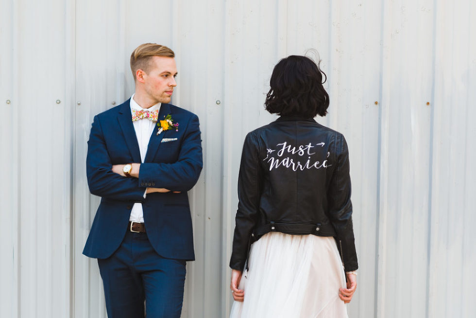 bride wearing black just married leather jacket over white wedding dress next to groom in front of white metal wall Toronto wedding photography