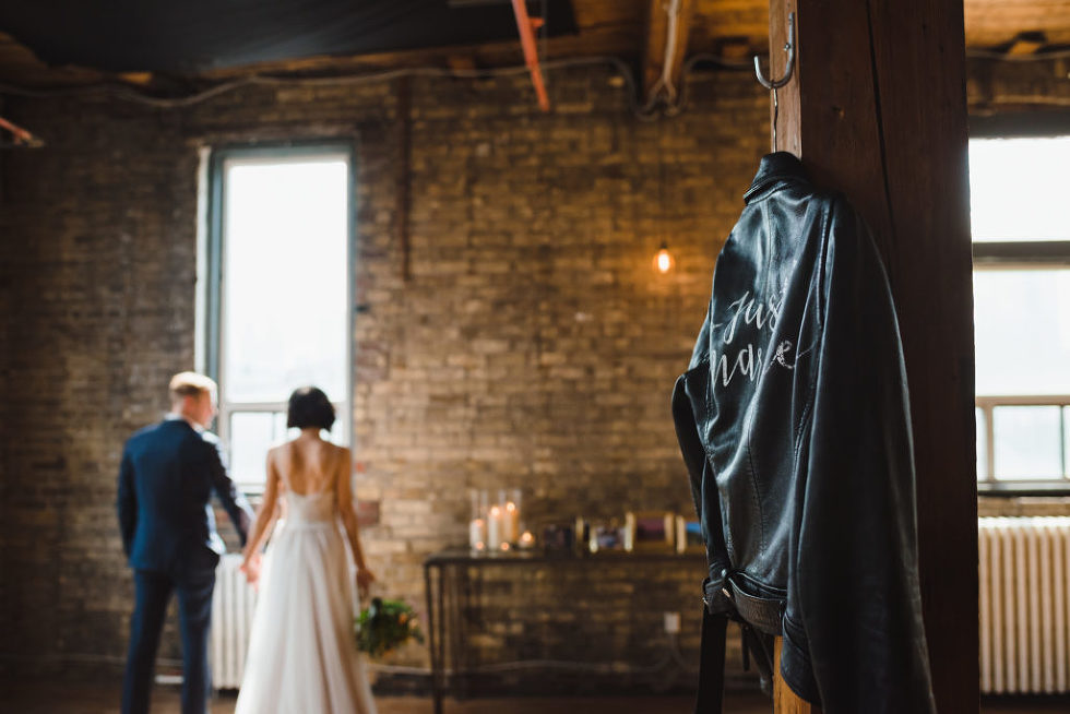 just married leather jacket hanging up on a wooden post with the bride and groom holding hands and facing brick wall and wedding decorations at the Jam Factory Toronto wedding photography