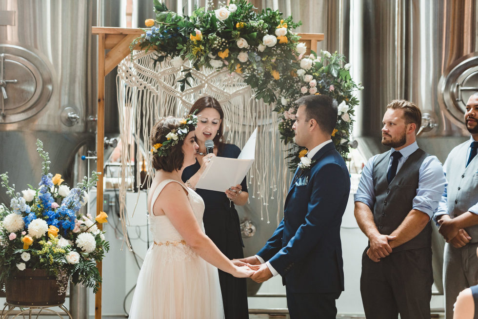 bride and groom holding hands as officiant performs wedding ceremony with brewing tanks in the background Toronto Junction Craft Brewing