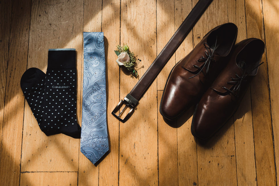 Grooms brown shoes, brown belt, blue necktie, and black and white polkadot socks laid out on wooden floor before wedding at La Paletta Mansion Burlington Ontario