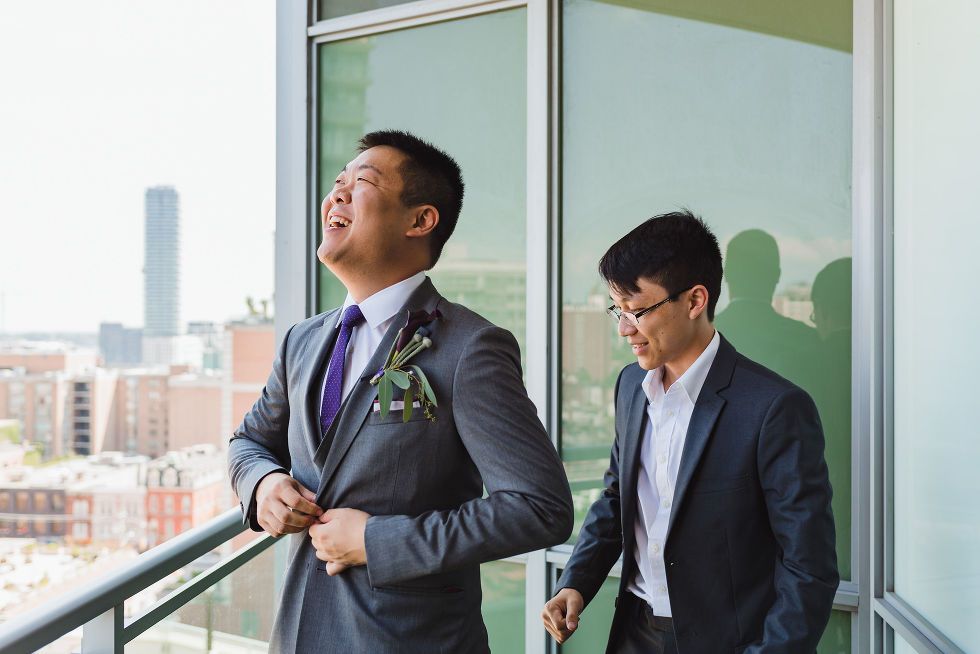 Groom laughing while he buttons up his suit in front of groomsman on a condo balcony before his Parisian inspired wedding at La Maquette in Toronto Ontario 