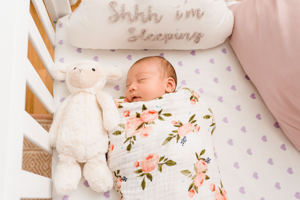 A baby swaddled in a floral blanket and sleeping in a crib with stuffed bunny and a pillow that says shhh i